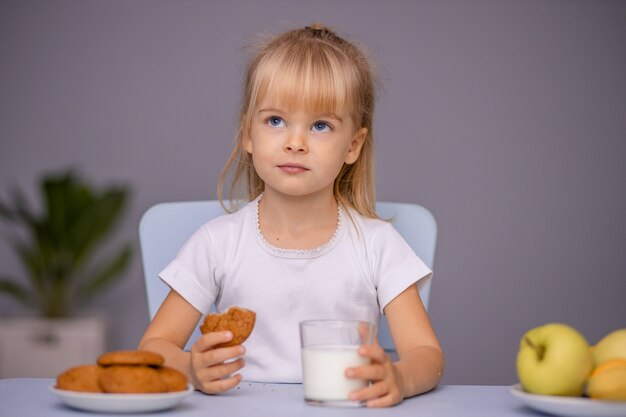 Menina bonitinha comendo biscoitos e bebendo leite em casa ou no jardim de infância