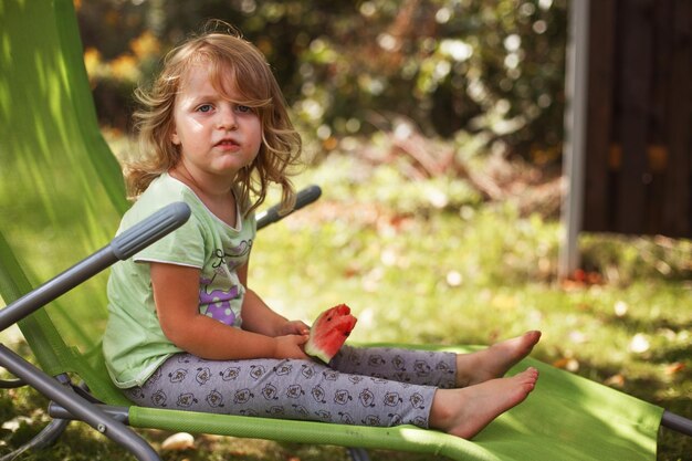 Menina bonitinha comendo melancia em uma espreguiçadeira no jardim no verão