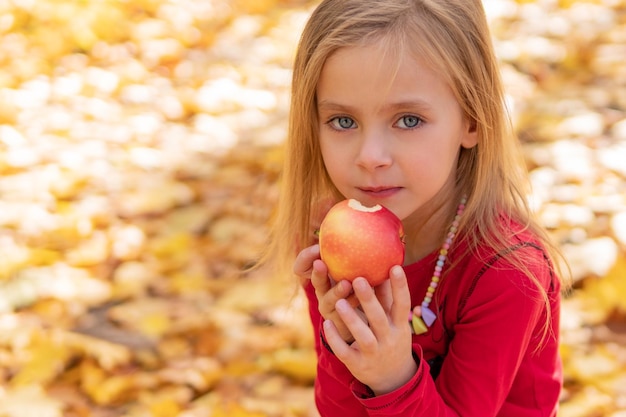 menina bonitinha comendo uma grande maçã vermelha em um fundo de folhagem de outono