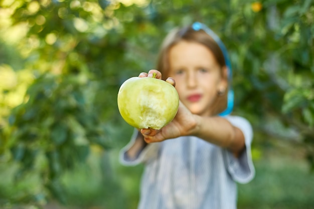 Menina bonitinha comer maçã verde no jardim doméstico ao ar livre, criança feliz, fazer caretas, colheita de outono, macieira, estilo de vida de comida vegetariana de dieta de nutrição saudável.