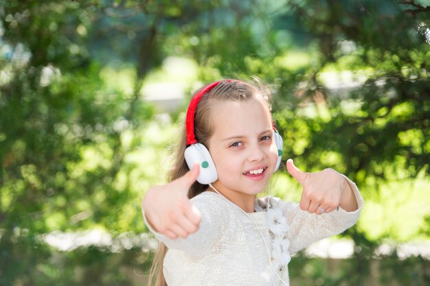Menina bonitinha curtindo música usando fones de ouvido no dia de verão e mostrar sim gesto