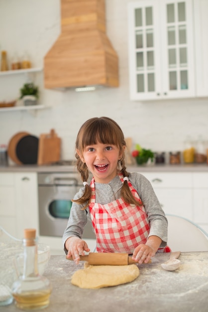 Menina bonitinha de 4 anos com rabo de cavalo desenrola a massa com um rolo na mesa da cozinha.