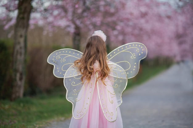 Menina bonitinha de cabelos compridos encaracolados em linda fantasia de fada rosa com asas grandes no jardim de sakura florescendo férias de primavera