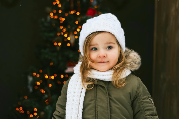 menina bonitinha de chapéu branco tricotado fica na varanda da casa de campo e decora a árvore de Natal para