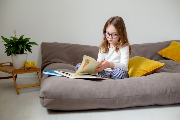 menina bonitinha de óculos, jeans e gola alta, está lendo um livro sentada na cama