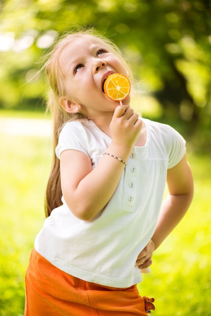 Menina bonitinha de pé na grama verde no parque e segurando pirulito laranja na mão.