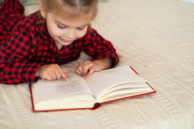 menina bonitinha de pijama vermelho de natal deitada na cama lendo livro