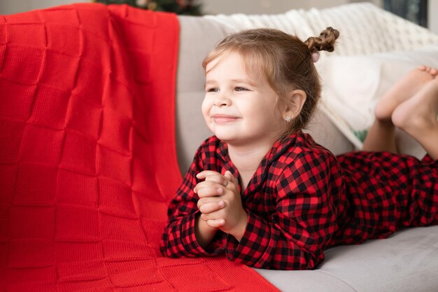 menina bonitinha de pijama vermelho de natal sorrindo deitada no sofá