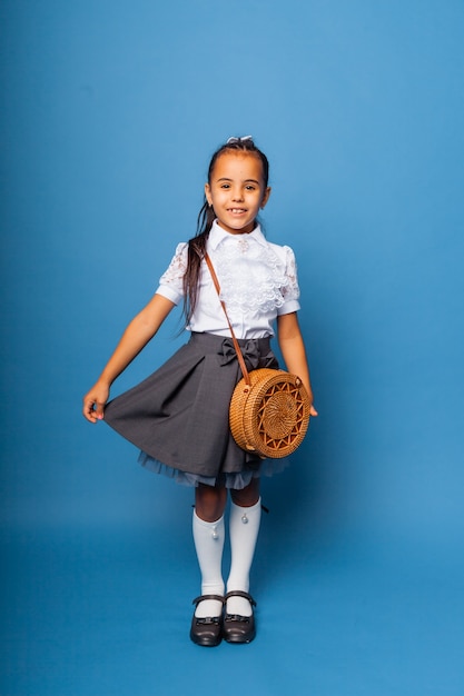 Menina bonitinha de sete anos, posando em uniforme escolar e segurando uma sacola redonda de vime sobre um fundo azul