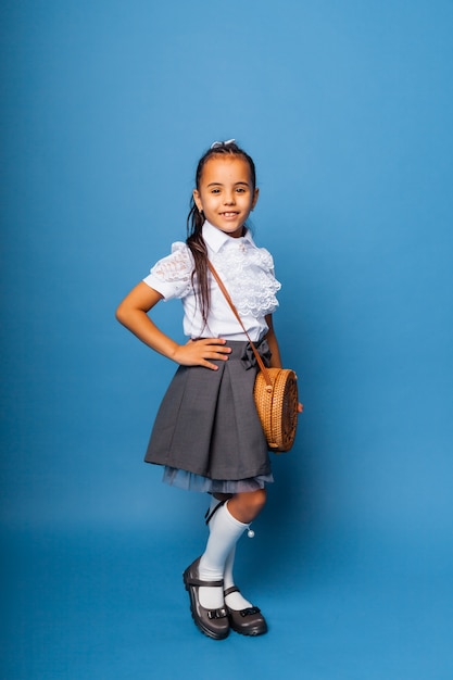 Menina bonitinha de sete anos, posando em uniforme escolar e segurando uma sacola redonda de vime sobre um fundo azul