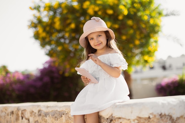 Menina bonitinha de vestido branco e chapéu de palha olhando para a câmera enquanto estiver usando smartphone