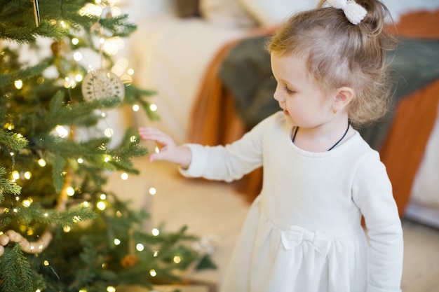 Menina bonitinha de vestido branco fica perto da árvore de Natal com decorações e luzes
