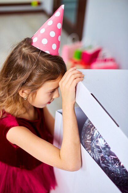 Menina bonitinha de vestido rosa e chapéu abre uma grande caixa de presente