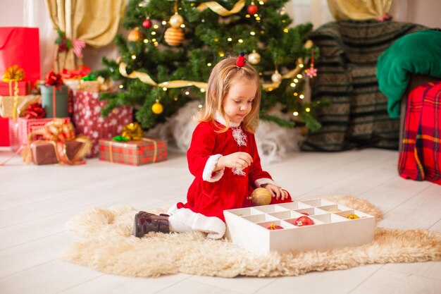 Menina bonitinha decorando uma árvore de Natal com enfeites coloridos em caixa de madeira Boas festas de Natal
