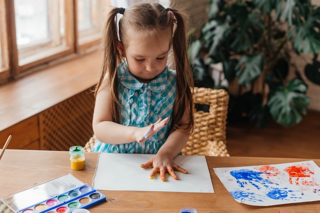 Menina bonitinha desenha com tintas e mãos. Palmas das crianças pintadas