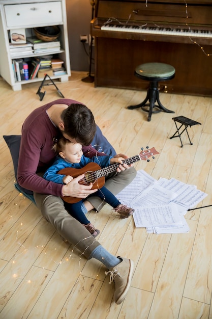 Foto menina bonitinha e seu pai bonito estão tocando violão e sorrindo enquanto está sentado na sala