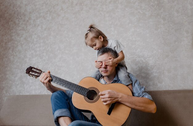 Menina bonitinha e seu pai bonito tocam guitarra e sorriem, sentado no sofá em casa. dia dos pais. cuidado e educação das crianças.