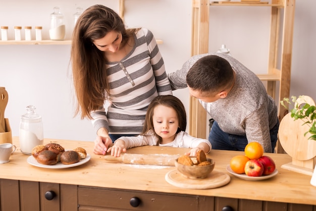 Menina bonitinha e seus lindos pais preparam comida e sorriam enquanto cozinha na cozinha em casa. amasse a massa em panquecas e pãezinhos