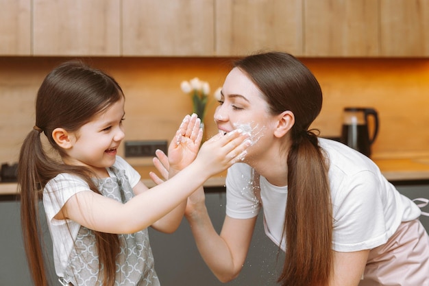 Menina bonitinha e sua jovem mãe de avental estão brincando com farinha e rindo enquanto amassam a massa na cozinha