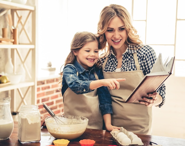 Menina bonitinha e sua linda mãe de avental estão lendo receita e sorrindo enquanto amassa a massa na cozinha