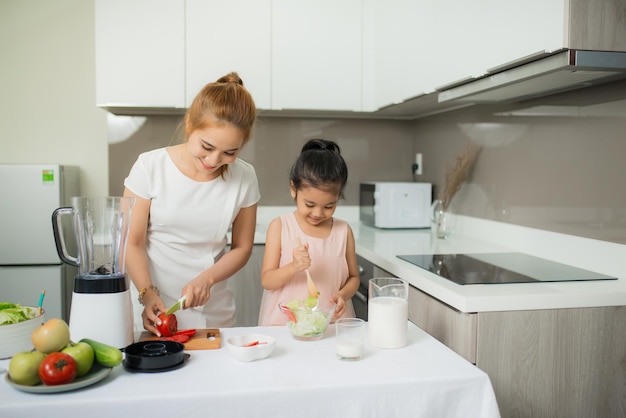 Menina bonitinha e sua mãe com chapéus de chef estão cortando legumes, cozinhando uma salada e sorrindo