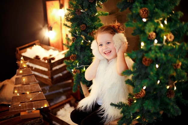 Menina bonitinha em abafador de pele sentado perto de árvore de natal