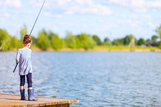 Menina bonitinha em botas de borracha, pesca do cais de madeira em um lago.