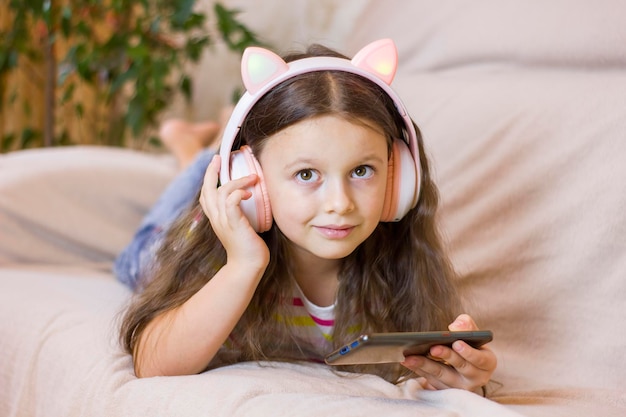 Menina bonitinha em fones de ouvido usando um smartphone ouve música sorrindo e deitada no sofá em casa