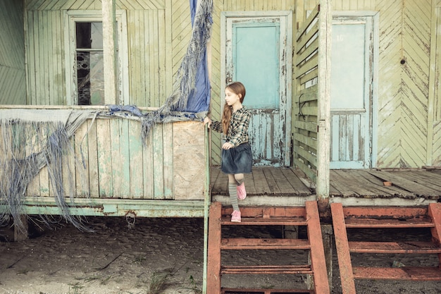Menina bonitinha em pé na escada da casa de madeira abandonada