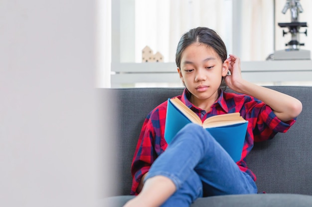 Menina bonitinha em roupas casuais lendo um livro deitado em um sofá na sala de estar