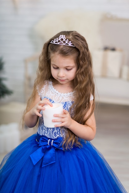 Menina bonitinha em um magnífico vestido azul com uma vela artificial nas mãos para a decoração da casa nas férias de Natal. Cenário para crianças.