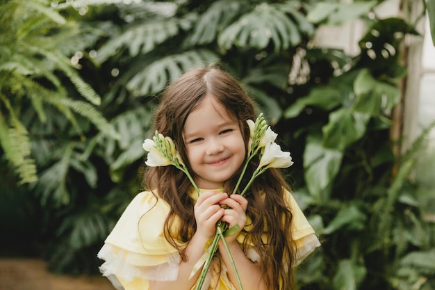 Menina bonitinha em um vestido amarelo segurando flores de primavera em suas mãos em pé contra o fundo das folhas