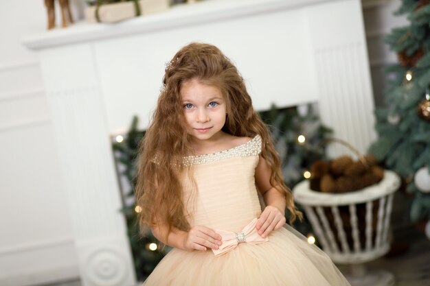 Menina bonitinha em um vestido bege exuberante posando perto da árvore de natal