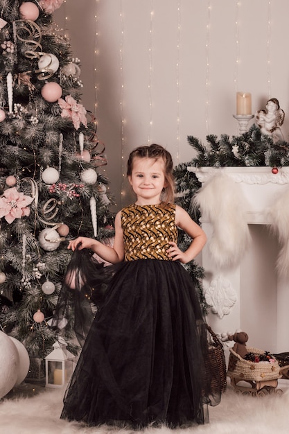 Menina bonitinha em um vestido preto e dourado perto de uma lareira e uma árvore de natal