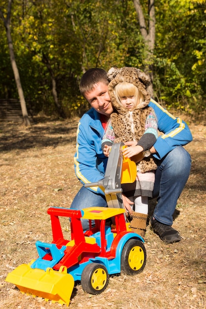 Foto menina bonitinha em uma roupa de gato malhado posando nos braços de seu pai ajoelhado ao ar livre no q park com um grande brinquedo de construção de plástico colorido com um carregador frontal