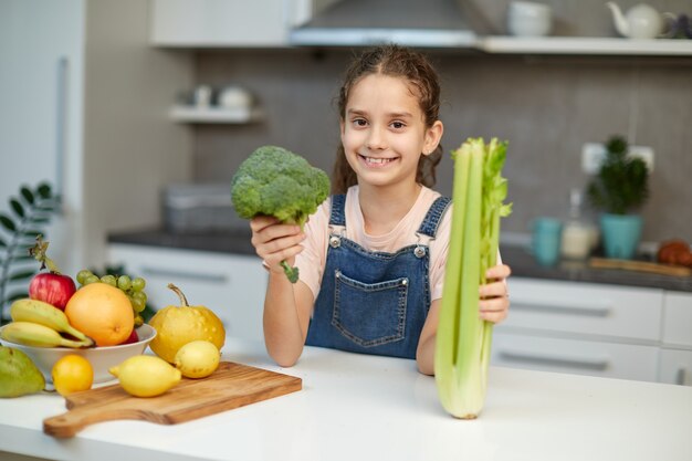 Menina bonitinha encaracolada fica perto da mesa na cozinha, segurando brócolis verde e aipo nas mãos.