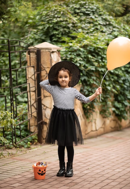 Menina bonitinha fantasiada de bruxa segurando balão Estande próximo balde de abóbora jackolantern com doces e doces Travessuras ou travessuras infantis no feriado de Halloween