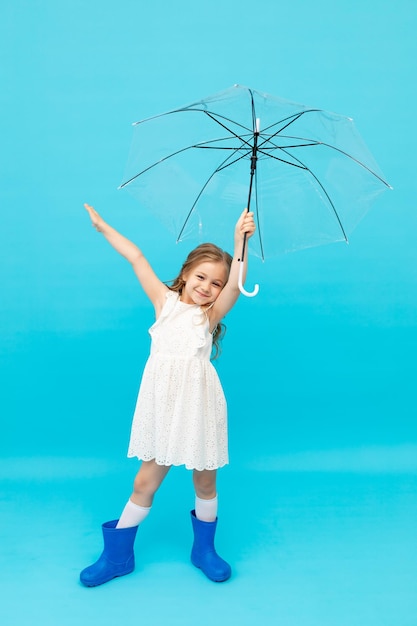 Menina bonitinha feliz em botas de borracha azul e um vestido branco de algodão segurando um guarda-chuva em um fundo azul no estúdio e sorrindo e brincando com espaço para texto