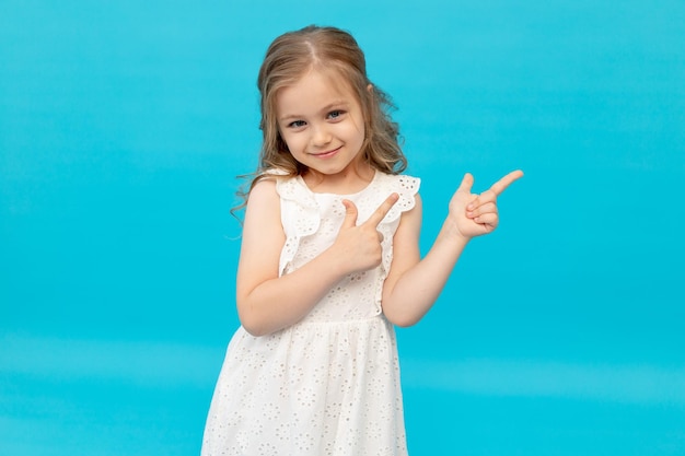 Menina bonitinha feliz em um vestido branco de algodão em um fundo azul no estúdio rindo mostrando os dedos no ver sorrindo e brincando um lugar para texto