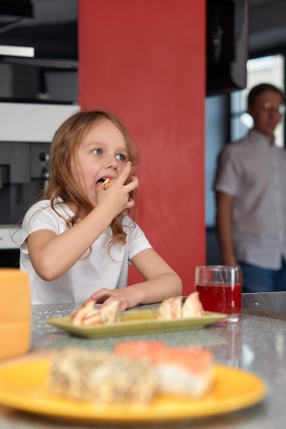 Menina bonitinha gosta de comer sushi Garota pré-escolar loira caucasiana alegre e engraçada com rolo de sushi e no fundo da cozinha