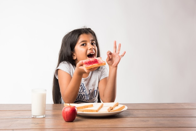 Menina bonitinha indiana ou asiática tomando café da manhã. Mesa cheia de fatias de pão com geléia, copo de leite, maçã fresca e ovo cozido