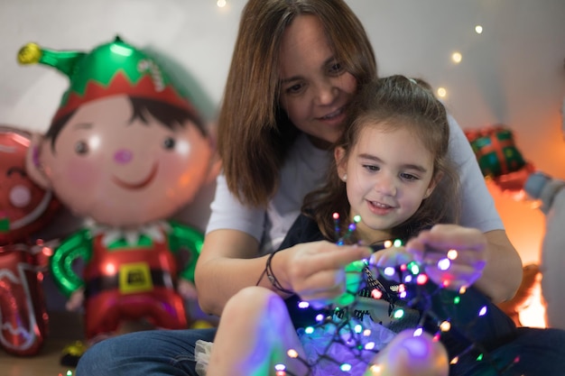 Menina bonitinha interagindo com interesse com as luzes de Natal sentada no colo da mãe que a ensina