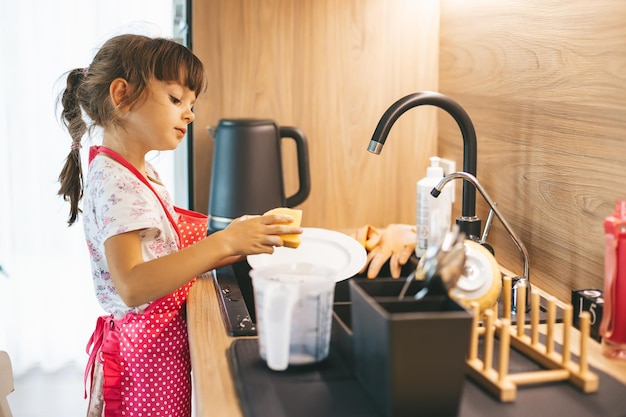 Menina bonitinha lavando pratos com esponja na pia da cozinha em casa