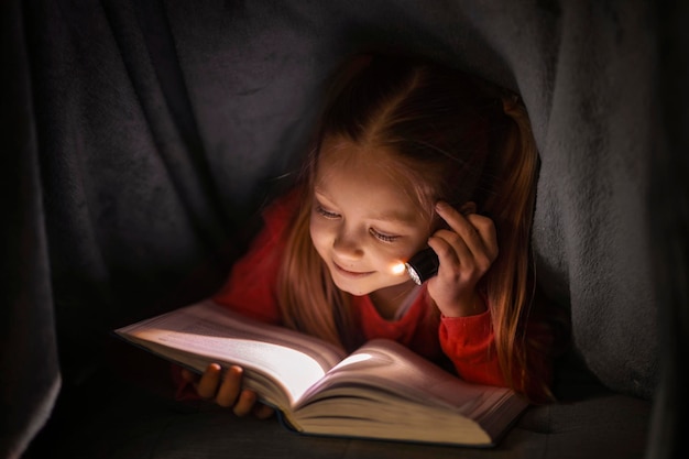 Menina bonitinha lendo livro enquanto se esconde sob o cobertor com lanterna