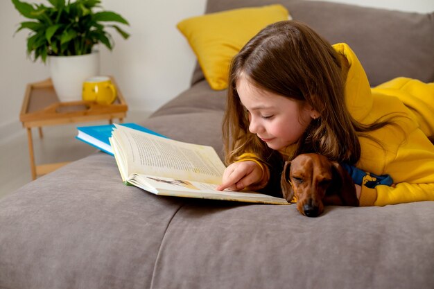 Menina bonitinha lendo um livro deitada na cama com um dachshund anão educando em casa