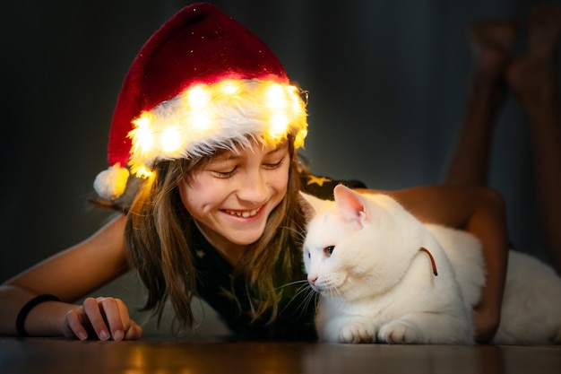 Menina bonitinha no chapéu de Natal segurando gato branco e sorrindo.