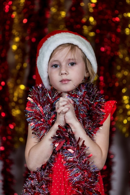 Menina bonitinha no chapéu de Papai Noel e com enfeites em volta do pescoço, fazendo desejo de presentes de Natal.