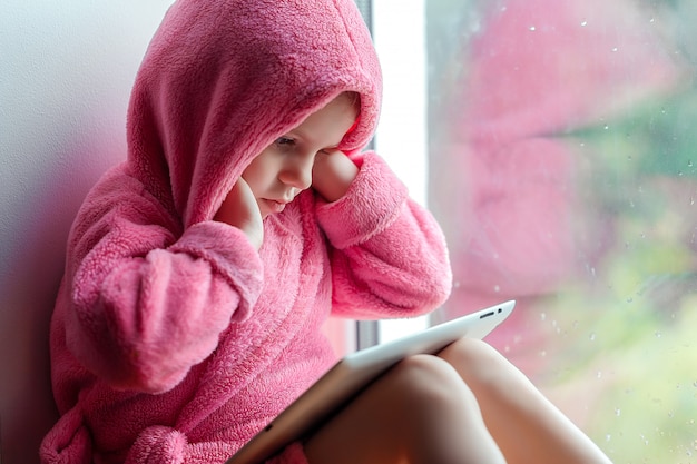 Menina bonitinha no roupão rosa usando o tablet pc enquanto está sentado no parapeito da janela.