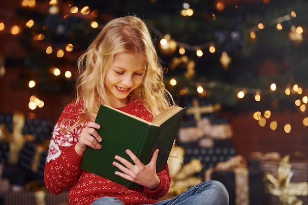 Menina bonitinha no suéter festivo vermelho lendo livro dentro de casa na época do feriado de Natal.