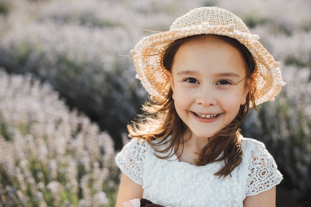 Menina bonitinha rindo contra um campo de flores, usando chapéu e camisa branca.